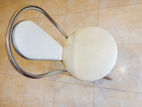 Chair with metal legs and frame, circle seat and back-rest padded with white faux leather stands on the ceramic tile floor, top view in selective focus