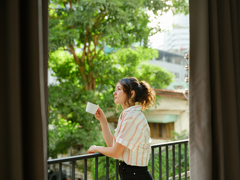 Cheerful woman drinking coffee  on the balcony in summer with trees visible from the window