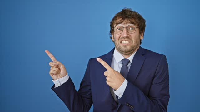 Shocked young man in business suit, nervously pointing aside - display of worry, surprise, and concern on an isolated blue background.