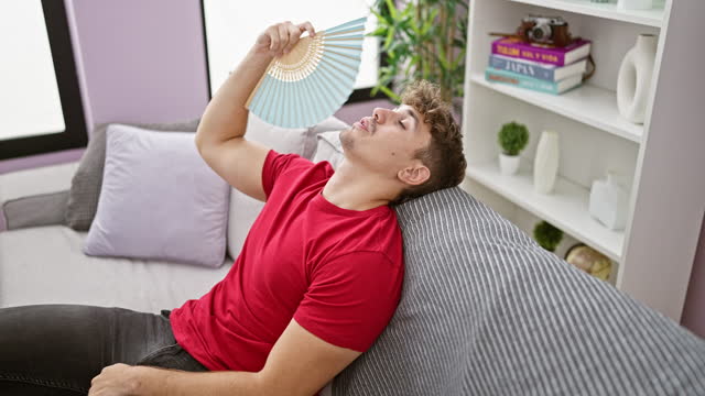 Hot weather woes, young hispanic man suffers, fanning himself with handfan sitting on sofa at home, exposing the struggles of indoor heat