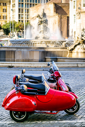 Rome, Italy, November 11 -- Some lovely and characteristic Vespa sidecar scooters parked in Piazza della Repubblica on the edge of the Rione Monti (Monti district) formerly called the Suburra district, in the historic heart of Rome. In the background, the famous Fountain of the Naiads, built between 1870 and 1912. Designed starting in 1946 by the Italian motorcycle brand Piaggio, the Vespa has become one of the ever-fashionable symbols of industrial culture and Italian design in the world. In 1980 the historic center of Rome was declared a World Heritage Site by Unesco. Image in high definition format.