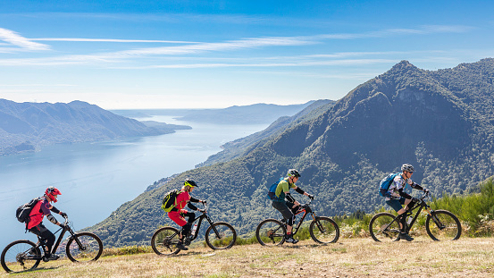 A group of experienced mountainbiker is riding in a row on an alpine pasture in front of scenic Lago Magiore.\nCanon EOS 5D Mark IV, 1/1000, f/8, 29 mm.