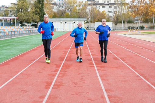Three senior Caucasian athletes in blue gear jogging on a red athletic track