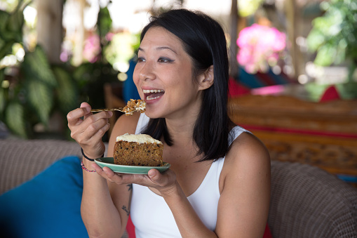 Close-up shot of Asian woman eating a slice of carrot cake with cream cheese icing. She's sitting alone in a cafe
