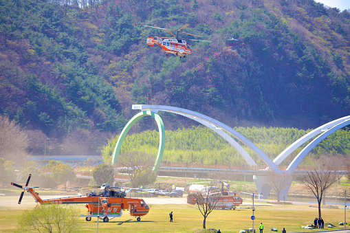 Ulsan, South Korea - June 21, 2023: A dynamic scene at Taehwa Park with one firefighting helicopter soaring in the air and two more stationed on the ground, showcasing coordinated emergency response efforts.