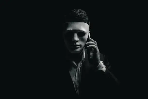 Unknown businessman wearing mask with covered face using mobile phone makes an anonymous call intimidating and threatening the interlocutor on dark background. hacker callcenter concept.