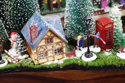 Stock photo showing a beautiful Christmas village display, which features numerous illuminated houses. The houses have been placed on artificial grass and sprinkled with fake snow to create a snowy scene, on top of a chest of drawers, complete with a forest of plastic spruce trees.