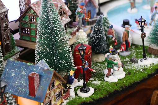 Stock photo showing a beautiful Christmas village display, which features numerous illuminated houses. The houses have been placed on artificial grass and sprinkled with fake snow to create a snowy scene, on top of a wooden cabinet, complete with a forest of plastic spruce trees.