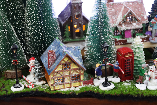 Stock photo showing a beautiful Christmas village display, which features numerous illuminated houses. The houses have been placed on artificial grass and sprinkled with fake snow to create a snowy scene, on top of a chest of drawers, complete with a forest of plastic spruce trees.