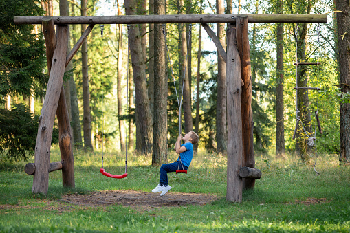 A little boy sits on a large rope swing in a sunny forest. The child plays with the ropes, twisting them and then quickly spins around its axis.