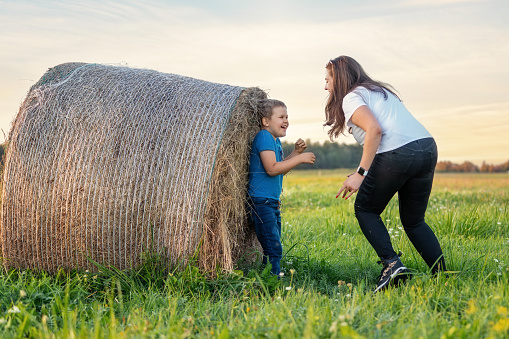 A mother has discovered and catches her son hiding behind a big hay bale. Fun outdoor games in the village during summer vacation with a child.