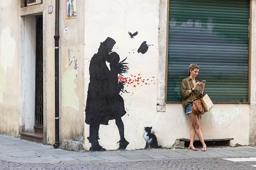 Padua, Veneto, Italy - Jun 22nd, 2023: Woman leaning against wall in the street using smartphone by a Kenny Random's romantic graffiti on a corner of Padova city center