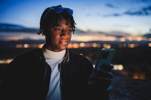 Young adult with a smartphone during a dusk, with the blurry city lights visible behind them. Concept of technological lifestyle.