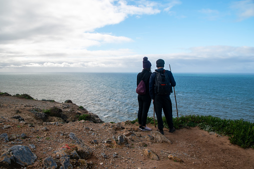 A young tourist couple, standing on a rugged cliff overlooking the vast Atlantic Ocean, gazes in awe at the untamed beauty of nature during their adventurous hike.