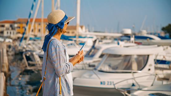 With the timeless beauty of Novigrad town in Croatia as the backdrop,a side view captures a female tourist holding her smart phone while standing at the charming harbor. A seamless fusion of modern connectivity and coastal charm