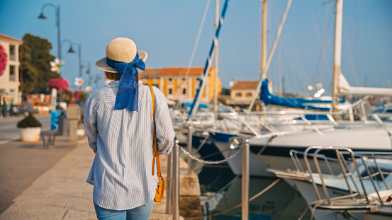 In the enchanting harbor of Novigrad town,Croatia,a rear view captures the grace of a female tourist,adorned with a sun hat,strolling amidst the coastal charm