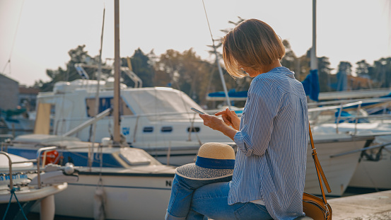 Along the tranquil pier in Novigrad,Croatia,a side view captures a female tourist immersed in the beauty of the moment,using her smart phone. A blend of modern connectivity and the timeless charm of coastal exploration