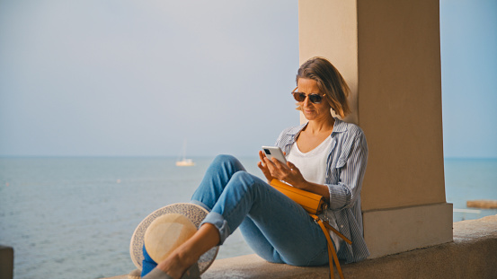 In the picturesque colonnade by the sea in Novigrad,Croatia,a female tourist indulges in the beauty of the moment,using her smart phone while seated on the retaining wall. A seamless blend of modern exploration and coastal serenity