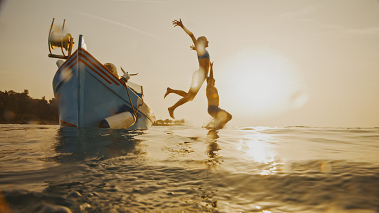 Embrace the freedom of the open sea. In a side view shot,a carefree couple jumps into the vast seascape from a motorboat against a clear sunset sky,a scene of pure joy and adventurous spirits amid the beauty of the setting sun