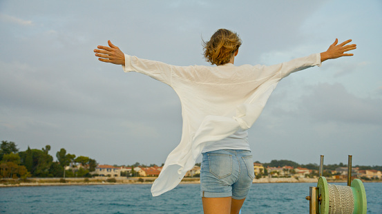 Embracing the carefree spirit of travel,a female tourist stands on a boat in the sea,arms outstretched,wearing a flowing white shirt against the vast sky. This scene encapsulates the joy of exploration and connection with nature