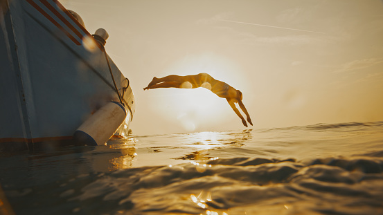 Embrace the freedom of the open water. In a full length side view,a backlit woman gracefully dives into the sea from a motorboat against the radiant sunset sky,an empowering silhouette against the canvas of the evening