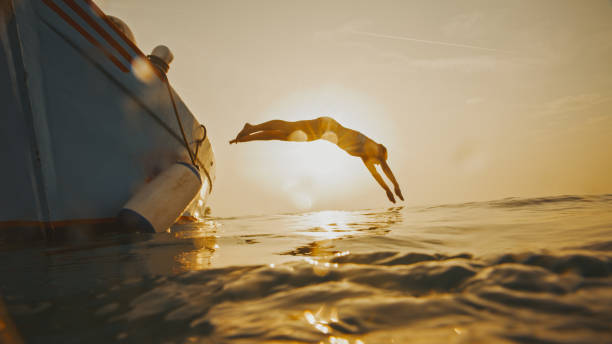 Full length side view of back lit woman diving into sea from motorboat against sky during sunset Embrace the freedom of the open water. In a full length side view,a backlit woman gracefully dives into the sea from a motorboat against the radiant sunset sky,an empowering silhouette against the canvas of the evening just do it stock pictures, royalty-free photos & images