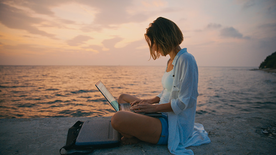In the tranquil ambiance of Istria,Croatia,a side view frames a focused individual working on their laptop while seated on a retaining wall by the sea. The sunset sky serves as a picturesque backdrop,blending productivity with the soothing beauty of the coastal evening.