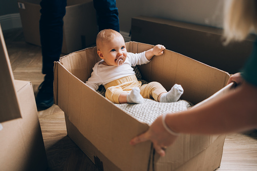 As the family settles into a new home, witness the endearing sight of a baby in a cardboard box, turning the moving day into a playful exploration, embraced by the love of her parents.