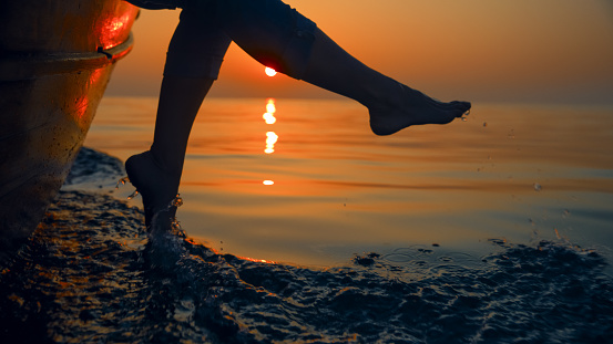 Silhouetted against the stunning Istrian sunset in Croatia,a woman's low section joyfully splashes sea water with her feet from a boat. The captivating scene blends the beauty of nature with the carefree spirit of a coastal moment