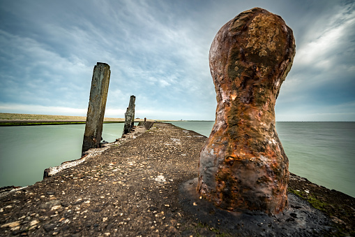 Rusted bollard on a ship mooring with a round concrete pier and a view over the sea and the cloudy sky\nFlaauwers Haven, Moriaanshoofd, Schouwen Duiveland, Oosterschelde, Zeeland, Netherlands, Europe
