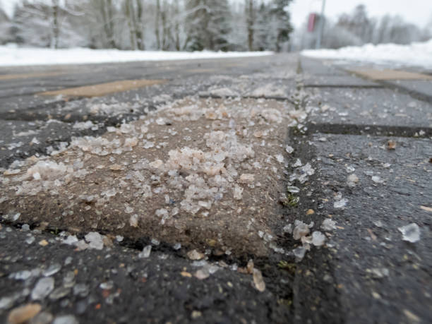 Salt grains on sidewalk surface in the winter. Applying salt to keep road clear from ice or snow in winter. Macro view of salt grains with white winter scenery in bacground
