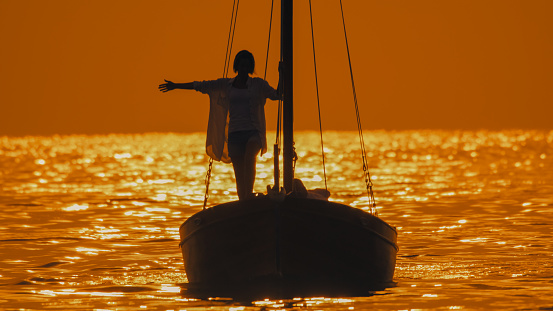 Embracing the idyllic sunset in Istria,Croatia,a carefree woman stands with her arm outstretched on the silhouette of a sailboat in the enchanting seascape. The scene captures the serene beauty of the Adriatic coast,where the sun sets in hues of warmth