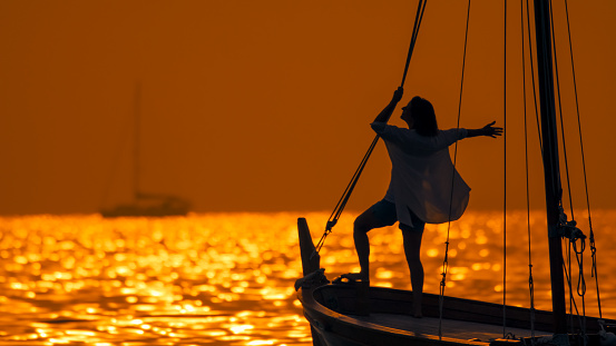 Against the enchanting backdrop of Istria's sunset,a carefree woman stands with her arm outstretched on the silhouette of a sailboat. The scene captures the idyllic beauty of the Adriatic Sea,blending the warmth of the setting sun with the tranquility of the coastal voyage in Croatia