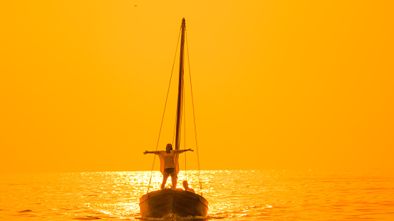 Serene silhouette,A woman stands on a sailboat in the sea,arms outstretched,against the golden reflections of the sunset in Istria,Croatia. This captivating scene harmonizes the beauty of nature with the tranquility of the moment