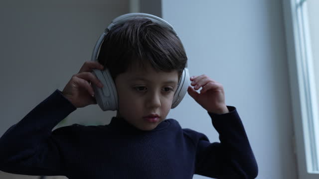 One small boy listening to music, song, or audio with headphones over the ear standing by window at home. Child wears noise cancelling headset