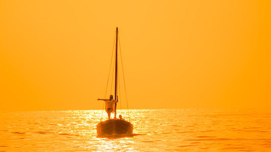 Amidst the golden reflections of the sunset in Istria,Croatia,a carefree woman stands with her arm outstretched on the silhouette of a sailboat,embracing the enchanting seascape
