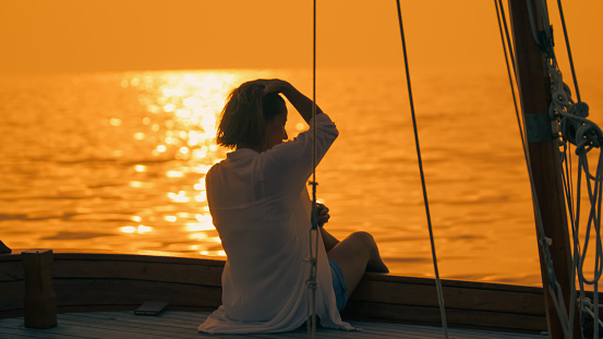 In the embrace of Istria's sunset in Croatia,a rear view captures a mid adult woman sitting on a sailboat,her hand gently in her hair. The scene reflects a tranquil moment of reflection,as the sun dips below the horizon,casting a warm glow over the Adriatic seascape