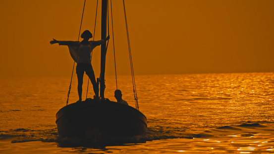 Embracing the carefree spirit of Istria's sunset in Croatia,a woman stands with her arm outstretched on the edge of a sailboat,with a man sitting behind in the idyllic seascape. The scene captures the joy and serenity of a shared moment,where the Adriatic unfolds its enchanting beauty under the canvas of the setting sun