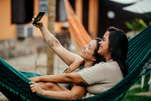 Young lesbian couple smiling and taking selfies while lying together in a hammock outside in a back yard