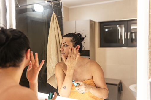 Woman applying facial cream in a bathroom, looking in the mirror