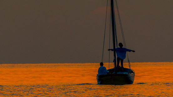 Embracing the warmth of Istria's idyllic sunset in Croatia,a woman stands with arms outstretched on a sailboat,with a man sitting behind in the vast orange seascape