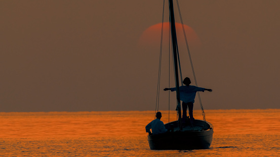 Against the backdrop of Istria's idyllic sunset in Croatia,a woman stands with arms outstretched on a sailboat,with a man sitting behind in the vast rippled seascape. The scene encapsulates a shared moment of joy and tranquility in the enchanting glow of the setting sun