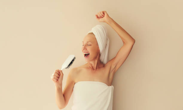 幸せで陽気な中年女性が髪を櫛で歌い、シャワーを浴びた後、濡れた髪を頭に巻いてタオルを頭に巻いて乾かした後、家で笑っています - hf7 ストックフォトと画像