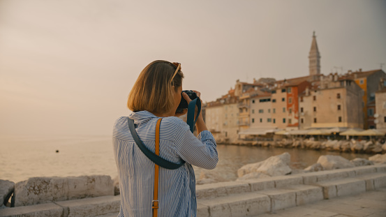 In the charming town of Rovinj,Croatia,a rear view captures a female tourist photographing the old town while walking on a pier by the sea. The scene encapsulates the picturesque beauty and cultural exploration along the Adriatic coast