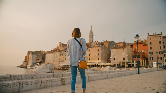 In the scenic town of Rovinj,Croatia,a rear view captures a female tourist walking on a pier by the sea,gazing at the captivating old town. The scene reflects the enchanting blend of coastal charm and historic architecture along the Adriatic coastline