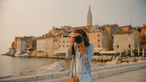 Capturing memories in the historic town of Rovinj,Croatia,a mid-adult female tourist photographs while looking at the camera against the backdrop of the charming old town. The scene reflects the joy of exploration and cultural appreciation during a vacation along the Adriatic coast