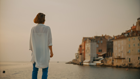 In the picturesque town of Rovinj,Croatia,a rear view captures a female tourist standing by the sea,looking at the captivating old town against the clear sky. The scene embodies the tranquil beauty and cultural richness of the Adriatic coastal setting