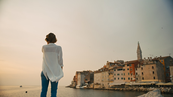 In the enchanting glow of sunset in Rovinj,Croatia,a rear view captures a woman standing against the clear sky and old town. The scene epitomizes the tranquil beauty of the Adriatic coastal evening,where the sun paints the horizon in warm and vibrant hues