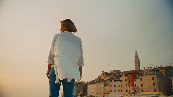In the contemplative ambiance of sunset in Rovinj,Croatia,a low-angle view captures a thoughtful woman standing against the clear sky and old town. The scene reflects the serene beauty of the Adriatic coastal evening,where the sun's warm hues paint the horizon