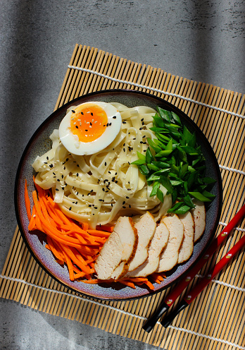 Traditional asian udon noodles with chicken, carrot, eggs, sesame and chives on dark background in sunlight. Served in a bowl. Top view. Copy space for text